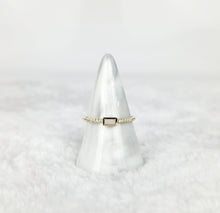 Charger l'image dans la galerie, Une Bague Livia de la fine joaillerie MONA présente une bande dorée avec une pierre centrale rectangulaire en pierre noire, flanquée de minuscules pierres claires. Il repose élégamment sur un support de bague en forme de cône marbré blanc et gris sur un fond blanc et pelucheux flou.