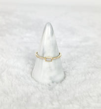 Charger l'image dans la galerie, Une bague en or avec une pierre centrale rectangulaire et de petits diamants est exposée sur un porte-bague en marbre blanc en forme de cône. Le fond est un tissu blanc doux et moelleux. Cette pièce exquise, nommée Bague Livia par MONA fine joaillerie, incarne l'élégance et le raffinement.