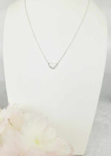 Charger l'image dans la galerie, Un délicat Collier Nourri-Source 10 mm, doté d'un pendentif cœur entrelacé et méticuleusement confectionné par MONA joaillerie fine, est exposé sur un support buste blanc. L'arrière-plan est légèrement flou et une grappe de fleurs rose tendre et blanches est visible dans le coin inférieur gauche de l'image.