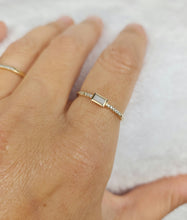 Charger l'image dans la galerie, Gros plan d'une main portant une délicate Bague Livia de joaillerie fine MONA avec une pierre centrale rectangulaire, entourée de petits diamants sertis le long de l'anneau. La main repose sur une surface douce et blanche, et une autre simple bande dorée est visible sur le doigt à côté.