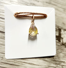 Load image into Gallery viewer, Le « Pendentif Goutte Éternelle » de la joaillerie fine MONA, doté d'un pendentif en forme de larme orné de pierres précieuses jaunes et claires, est élégamment suspendu à une chaîne tressée en or 10 carats. Cette pièce exquise est magnifiquement mise en valeur sur un fond carré blanc, reposant gracieusement sur une surface en bois.