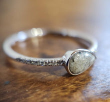 Charger l'image dans la galerie, Gros plan sur la Bague Goutte de vie (avec zircons) de MONA fine joaillerie, composée d'une pierre mouchetée en forme de goutte dans un chaton en argent et de petits zircons transparents sur la bande, reposant sur une surface en bois.