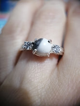 Charger l'image dans la galerie, Gros plan d'une main présentant une élégante Bague Trinité de la Bijouterie Langlois avec une pierre précieuse opaque centrale flanquée de pierres claires étincelantes sur un anneau délicat.