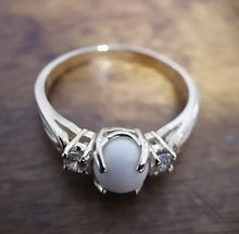 Charger l'image dans la galerie, Un gros plan d'une élégante Bague Trinité avec une pierre précieuse opaque centrale flanquée de petits détails cristallins sur une bande polie, exposée sur une surface en bois à la Bijouterie Langlois.