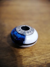 Charger l'image dans la galerie, Gros plan d'une toupie métallique colorée avec un arrière-plan flou, mettant en valeur un design bleu et argent vibrant avec des gravures numériques et des inclusions Bijoux La Précieuse autour du trou central.