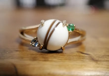 Charger l'image dans la galerie, Photographie en gros plan d'une élégante Bague Angélique de MONA fine joaillerie comportant une pierre centrale blanche avec des stries brunes, flanquée de petites pierres précieuses vertes et bleues, le tout serti sur une délicate bande dorée.
