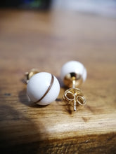 Charger l'image dans la galerie, Une paire de Perles d'oreille Boucle d'oreille sphériques blanches avec des accents dorés et un motif à bande unique, fabriquées à partir de Bijoux La Précieuse, reposant sur une surface en bois.