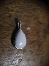 Charger l'image dans la galerie, Un pendentif Goutte15 avec une pierre de lait maternel lisse sertie dans un cadre métallique simple, reposant sur un fond en bois texturé par MONA fine joaillerie.