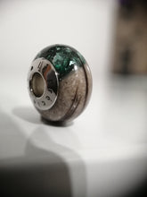 Charger l'image dans la galerie, Un gros plan d'une perle de verre MONA fine joaillerie Perle européenne Luxe avec un noyau métallique, mettant en valeur un mélange complexe de textures vertes et sableuses étincelantes, artistiquement mises en valeur sur un fond flou.