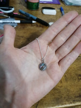 Charger l'image dans la galerie, Une main tenant un délicat Pendentif Griffes en acier inoxydable de Bijoux La Précieuse avec une chaîne et une pièce maîtresse en pierre multicolore scintillante, présentée sur fond de bureau parsemé de divers objets.