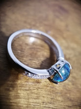 Charger l'image dans la galerie, Une bague Bague Précieuse de 8 mm de MONA fine joaillerie avec une pierre opalescente bleue scintillante, accentuée par une demi perle, sertie dans un design moderne, placée sur une surface en bois.