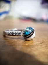 Charger l'image dans la galerie, Un gros plan d'une élégante Bague Précieuse fine joaillerie MONA 8 mm avec des zircons bleus chatoyants et de petits diamants sertis dans l'anneau, placés sur une surface en bois avec un fond légèrement flou.