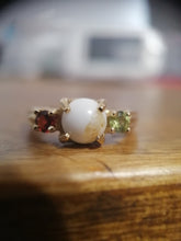 Charger l'image dans la galerie, Une élégante bague Bague Trinité avec une pierre centrale blanche flanquée de petites pierres précieuses multicolores, reposant sur une surface en bois avec un fond légèrement flou. Cette bague personnalisée est disponible chez Bijouterie Langlois.