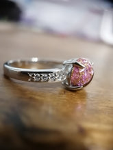 Charger l'image dans la galerie, Un gros plan d'une élégante bague en argent de joaillerie fine MONA avec une pierre cristalline rose sertie, complétée par de plus petits zircons transparents incrustés le long d'un côté de l'anneau, affichés sur une surface en bois.