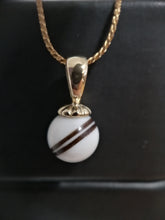 Charger l'image dans la galerie, Un élégant Pendentif RG150 de Bijoux La Précieuse avec une pierre sphérique blanche ornée d'une rayure centrale marron, suspendue à une fine chaîne dorée sur fond sombre, rappelle le lait maternel dans sa pureté et sa tranquillité.
