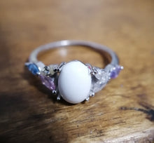 Charger l'image dans la galerie, Une élégante Bague Brielle de MONA fine joaillerie avec une pierre précieuse blanche proéminente, flanquée de petites pierres précieuses colorées, serties sur une surface en bois.