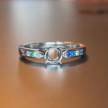 Charger l'image dans la galerie, Une bague Bague Précieuse de 5 mm avec une pierre précieuse centrale transparente flanquée d'un ensemble coloré de petites pierres précieuses sur un fond flou par MONA fine joaillerie.