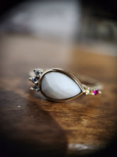 Charger l'image dans la galerie, Une élégante pierre précieuse en forme de larme sertie dans une bague en or, ornée de minuscules pierres précieuses sur une surface en bois chaude, dégageant un sentiment de beauté et de sophistication intemporelles. Cette Bague London de MONA fine joaillerie.
