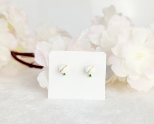 Les « Boucles d'oreilles Juri », une exquise paire de clous d'oreilles de la Bijouterie Langlois, sont ornées de petites pierres vertes et présentées sur une carte blanche. Le fond présente une ambiance douce et floue avec des fleurs aux couleurs claires, incarnant l'élégance douce caractéristique de la Bijouterie Langlois.