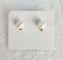 Charger l&#39;image dans la galerie, Gros plan sur les Boucles d&#39;oreilles Juri de la Bijouterie Langlois : des boucles d&#39;oreilles rondes blanches ornées d&#39;une ligne dorée diagonale et d&#39;une petite pierre verte au bas, élégamment présentées sur une carte carrée blanche. Parfaites pour celles qui recherchent des inclusions personnalisées dans leur collection de bijoux.
