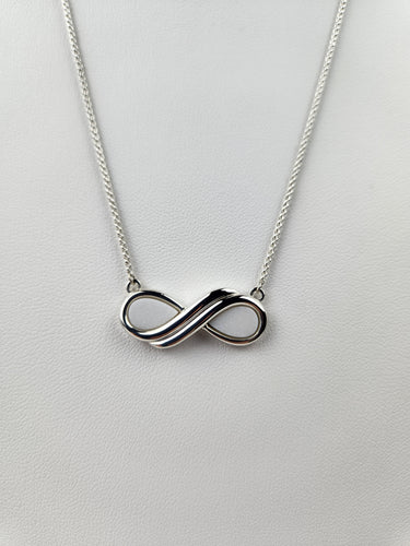 Un pendentif Collier Infinity en argent de MONA joaillerie fine sur une chaîne délicate repose sur un fond blanc, symbolisant l'éternité et l'amour éternel. Cette pièce offre une option d'inclusions personnelles.