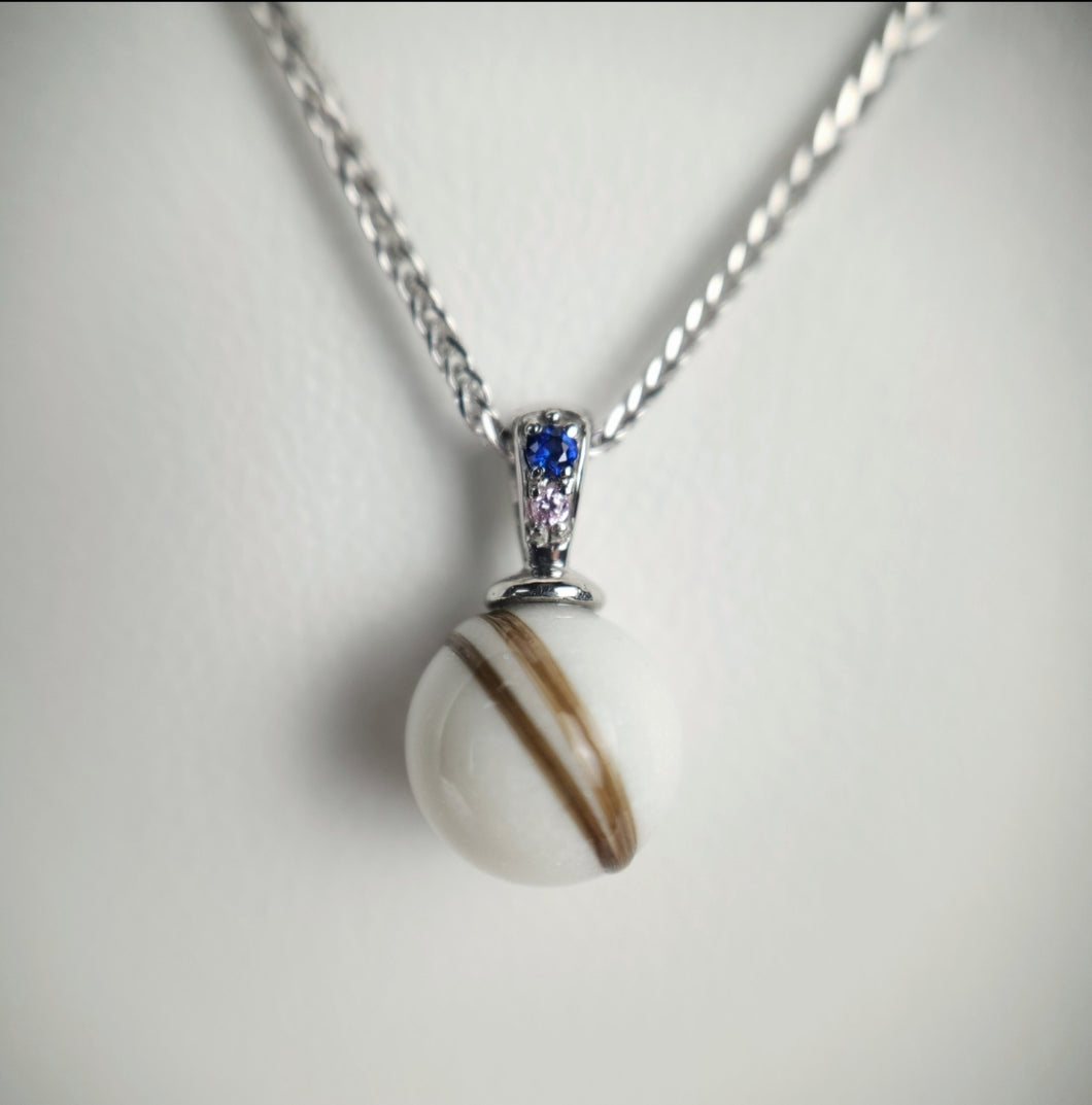 Un délicat Pendentif Double Zircon de MONA fine joaillerie comportant une perle aux stries brunes uniques suspendue à une chaîne en argent, accentuée par une petite pierre précieuse bleue étincelante au niveau de la bélière.
