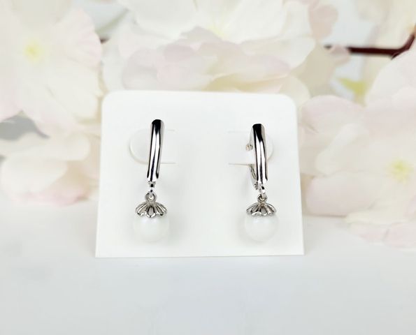 Une paire d'élégantes boucles d'oreilles Fermoirs en argent avec un design en forme de goutte d'eau, présentées sur un support blanc avec des accents floraux rose tendre en arrière-plan par Bijouterie Langlois.