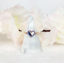 Charger l&#39;image dans la galerie, La Bague Coeur avec zircons de MONA fine joaillerie est une bague minimaliste en or avec un contour de cœur en pierres bleues, présenté sur un cône de marbre blanc et encadré par de douces fleurs rose pâle en arrière-plan.
