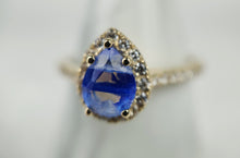 Charger l&#39;image dans la galerie, Gros plan de la Bague Bonheur Éternel de la Bijouterie Langlois, avec un anneau en or 10 carats avec une pierre centrale bleue en forme de goutte au centre, fixée par plusieurs griffes. La pierre précieuse est entourée d&#39;un halo de petits diamants étincelants. La bague est sertie sur un fond gris clair flou.
