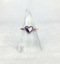 Charger l&#39;image dans la galerie, La Bague Coeur avec zircons de MONA fine joaillerie présente un motif en or en forme de cœur souligné de pierres bleues, élégamment présenté sur un support en marbre blanc sur un fond blanc doux.
