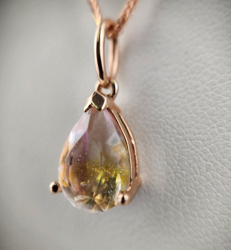 Un Pendentif Goutte Éternelle en forme de larme avec une monture en or fin MONA est suspendu à une chaîne en or fin 10 carats. La pierre précieuse présente un mélange de teintes jaunes et claires, créant un effet chatoyant sur le fond doux et neutre.