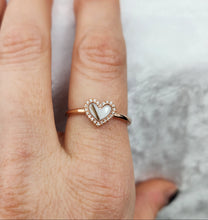 Charger l&#39;image dans la galerie, Une main porte la MONA fine joaillerie Bague Coeur avec zircons, une bague minimale avec un centre en forme de cœur et des zircons étincelants, sertis sur un fond clair doucement texturé.
