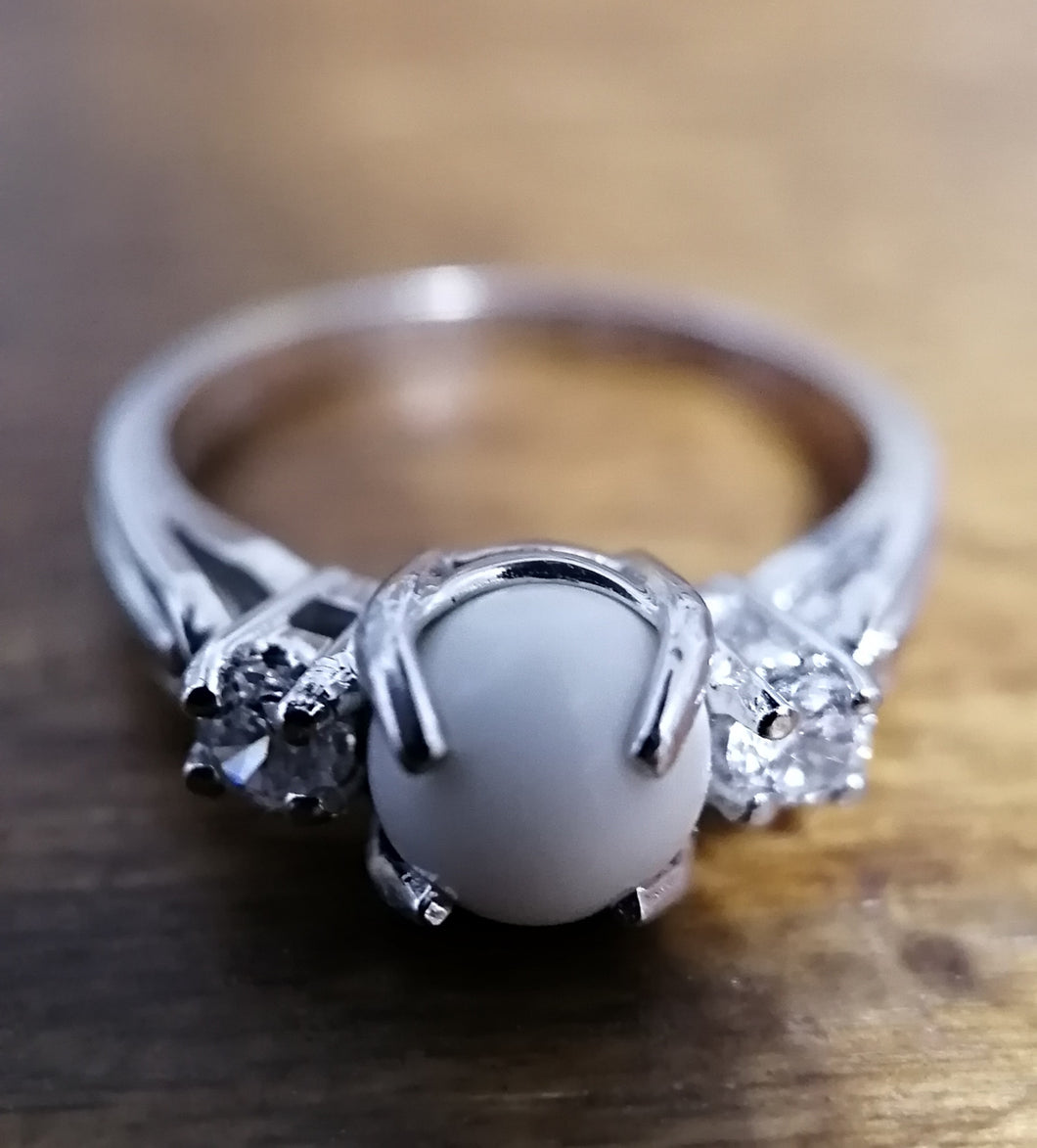 Une Bague Trinité personnalisable élégante en argent avec une perle centrale laiteuse flanquée de petits cristaux clairs, posée sur une surface en bois de la Bijouterie Langlois.