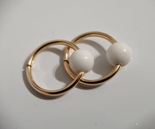 Une paire d'élégantes boucles d'oreilles créoles dorées avec des perles blanches ressemblant à des perles, inspirées des perles cordon ombilical, sur une surface blanche de MONA fine joaillerie.
